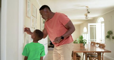 African american father measuring and marking child's height on hallway wall at home