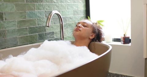 African american woman soaking in bubble bath in modern bathroom with green tile