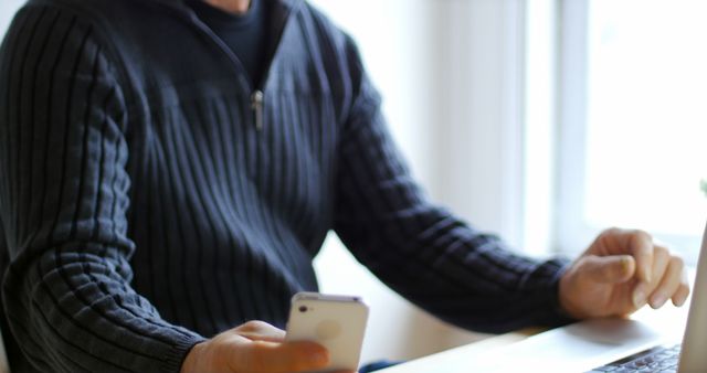 Man multitasking in modern workspace, using smartphone while working on laptop. Ideal for themes related to technology, modern work environments, and remote work.