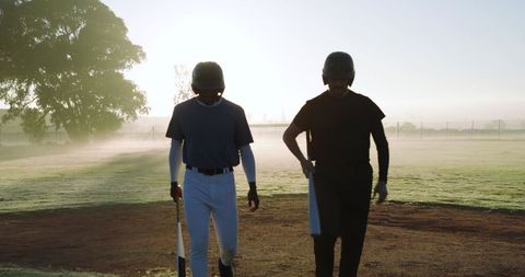 Backlit baseball teammates walking at sunrise carrying bats and helmets on misty diamond