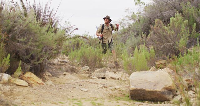 Man hiking on rocky desert trail surrounded by bushes and plants, holding fishing rods and carrying a backpack. Ideal for use in articles on outdoor adventures, travel blogs, nature programs, or promotional materials for hiking and fishing gear.
