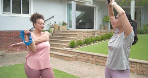 Two diverse women stretching with resistance band and hand weight on backyard patio