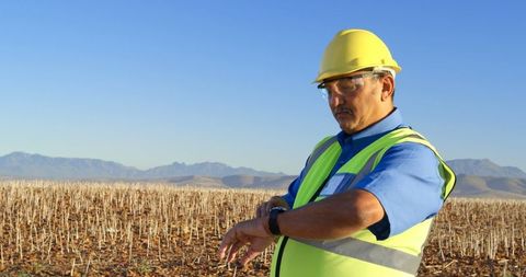 Engineer in Hard Hat Checking Smartwatch in Rural Landscape