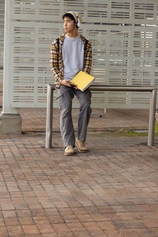 Asian commuter sitting on transit bench wearing headphones holding yellow folder