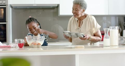 African american grandmother teaching granddaughter baking muffins and whisking in kitchen