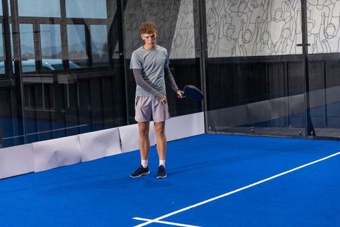 Young man standing on blue padel court holding racket beside glass wall mural and lines