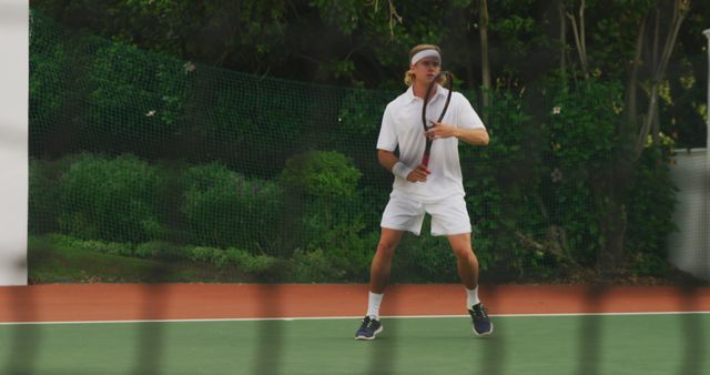 Male tennis player wearing white outfit and headband, holding racket, standing on outdoor court, preparing to receive serve, background of greenery and netting. Useful for sports articles, training guides, fitness content, promotional material for tournaments.