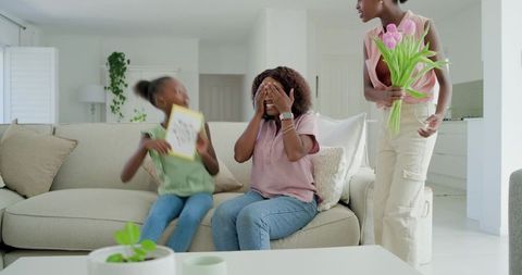 Woman covering eyes receiving surprise bouquet while child giving handmade card at home