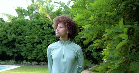 African american woman standing in mint pullover amid lush backyard greenery near pool