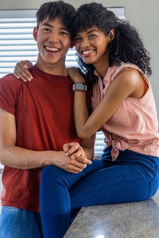 Young diverse couple smiling and embracing on kitchen counter showing smartwatch
