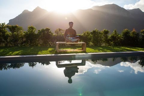 Asian man meditating on bench by reflective pool at sunrise with mountain backdrop serene