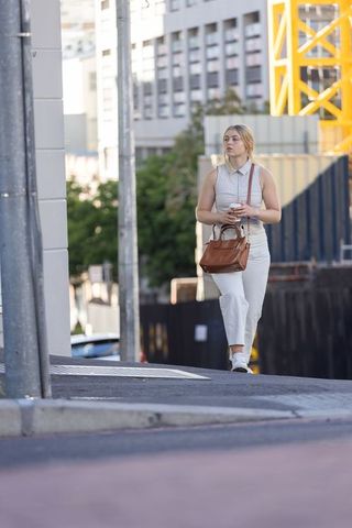 Walking with coffee and leather handbag past urban construction crane on sunny sidewalk
