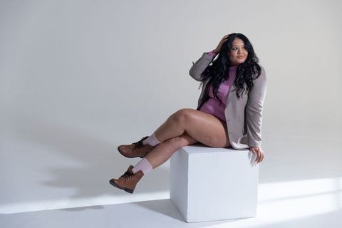 Curvy woman sitting on white cube wearing taupe blazer and pink bodysuit studio portrait