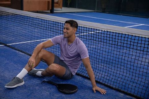 Young athlete resting by net on blue padel court holding paddle and ball, smiling