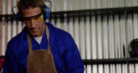 Welder inspecting metalwork at workbench wearing protective coveralls, leather apron, ear muffs