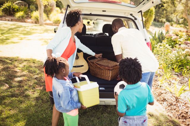 Family unpacking car for a picnic in the park. Parents and children taking out picnic basket, cooler, and guitar. Ideal for use in advertisements, family-oriented content, and articles about outdoor activities, family bonding, and leisure time.