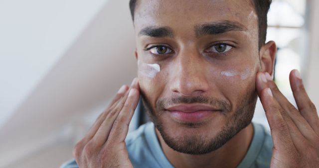 Young man applying face cream as part of daily skincare routine. Ideal for promoting beauty products, skincare tutorials, and advertisements focused on personal care and grooming for men.