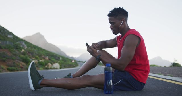 Young man sits on an asphalt road in a mountainous area, resting after physical activity. He concentrates on his smartphone while wearing earphones, athletic gear, and green sneakers. A water bottle is placed next to him, highlighting his dedication to fitness and hydration. This image is ideal for promoting fitness apps, sportswear brands, outdoor activity campaigns, and hydration products.