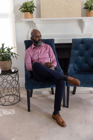 Mature african american man sitting in modern living room holding tumbler, relaxed