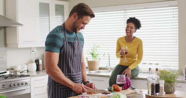 Middle-aged man in an apron chops vegetables on the kitchen counter while a woman sits on the countertop holding a glass of wine and laughing. Scene is full of warmth and relaxation. Perfect for articles and advertisements on home cooking, domestic life, relationship bonding, and lifestyle content.