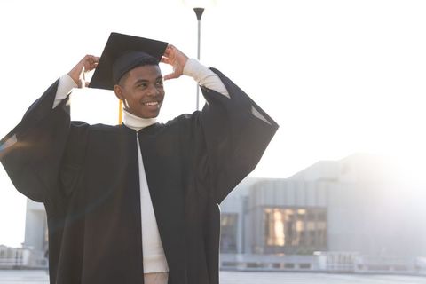 African american graduate smiling, adjusting mortarboard on rooftop with backlit lens flare