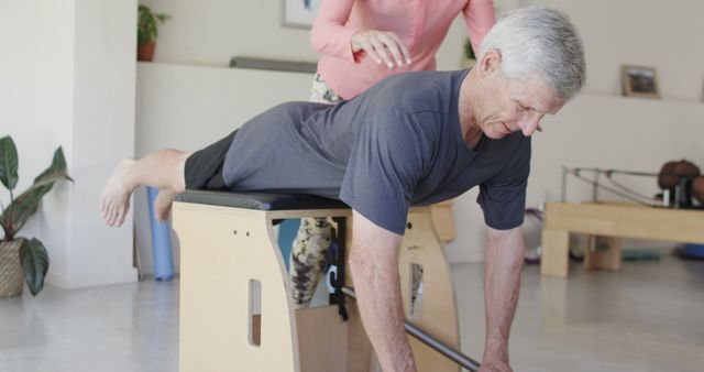 Senior man performing a Pilates exercise under the guidance of an instructor. Useful for illustrating themes related to fitness for seniors, physical rehabilitation, and guided exercise sessions. Can also be used to promote Pilates classes or physical therapy services targeting older adults.