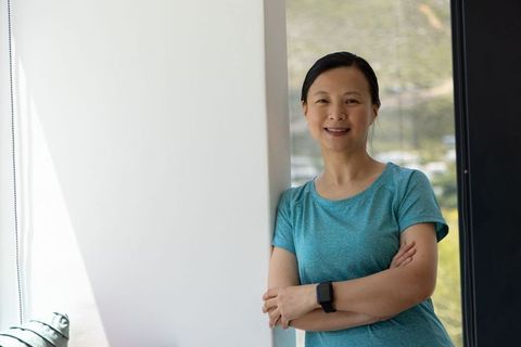 Mature asian woman leaning by window smiling wearing smartwatch and teal tee, modern interior