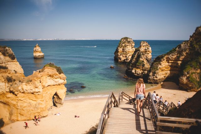 Beach scene with tourists walking down wooden stairs to sandy beach surrounded by impressive rocky formations in clear weather. Ideal for travel promotions, tourism websites, outdoor adventure blogs, vacation advertisements, and brochures showcasing scenic coastal landscapes.