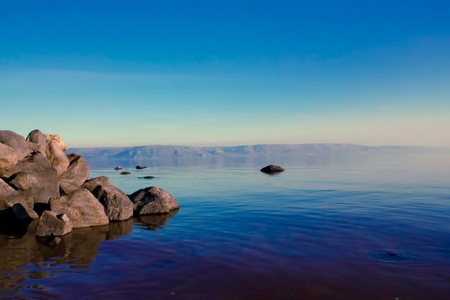 A peaceful lakeside scene with a rocky shore and calm water under a clear blue sky. Ideal for backgrounds, nature presentations, relaxation themes, travel websites, and promoting mindfulness or tranquility.