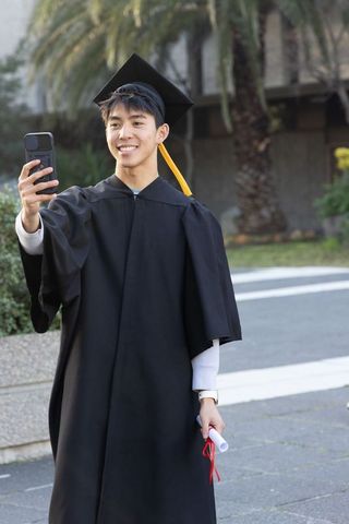 Asian graduate taking selfie on campus wearing cap and gown with yellow tassel holding diploma