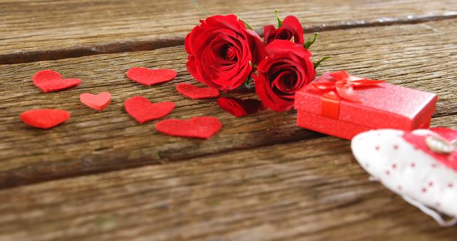 Red roses and a small gift box are accompanied by heart-shaped decorations on a wooden surface, with copy space. These elements typically symbolize romance and are often used in celebrations of love such as Valentine's Day or anniversaries.
