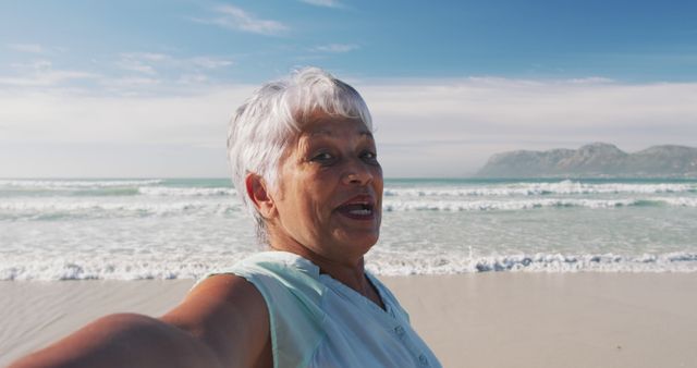Elderly woman taking a selfie at a beach with an ocean and mountains in the background. She is smiling and enjoying a bright sunny day, representing a fun and cheerful lifestyle during retirement. Useful for travel and vacation content, senior lifestyle blogs, and advertisements promoting leisure for the elderly.