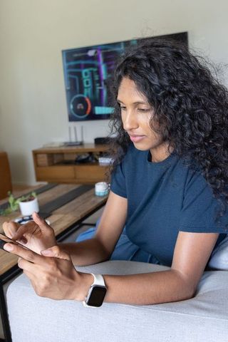 African american woman using smartphone and smartwatch relaxing on sofa in living room