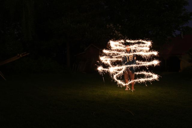 Person spinning sparklers in dark garden at night creating bright spiral light trails. Useful for images illustrating outdoor celebrations, festive activities, creative photography techniques, night-time fun, slow shutter photography, and recreational events.