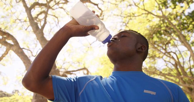 A young African American man is drinking water from a bottle outdoors, with copy space. Staying hydrated is essential for maintaining health, especially during physical activities.