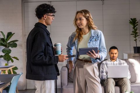 Diverse coworkers chatting in casual office lounge holding tablet and coffee
