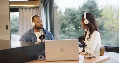 Diverse co-hosts recording podcast with laptop, microphones, headphones in sunlit studio