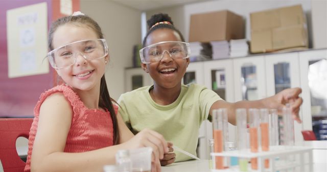 Two young girls of different ethnic backgrounds smiling and working on a science experiment in a classroom. They are wearing safety goggles and are surrounded by colorful test tubes and beakers. Useful for educational contexts, diversity in STEM promotion, teamwork, and hands-on learning activities.