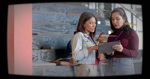 Two women collaborating over tablet at conference hallway, reviewing content with lanyards