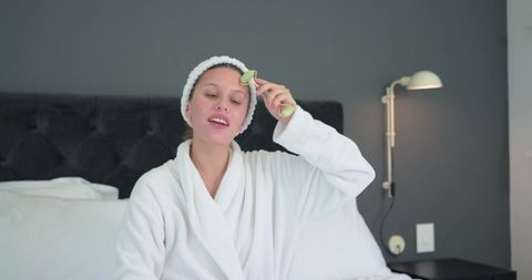 African american woman using jade roller wearing white robe for facial skincare routine