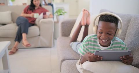 Smiling boy wearing headphones using tablet on sofa while woman holding coffee mug at home