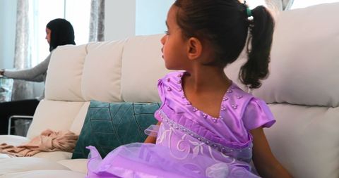 a young girl wearing a purple princess costume sits on a white couch in a modern living room. behind her, an adult is engaged in a task. this setting creates a warm and family-oriented atmosphere, ideal for illustrating a range of domestic lifestyles and activities. useful for content related to childhood fun, family lifestyles, home decor, and parenting.
