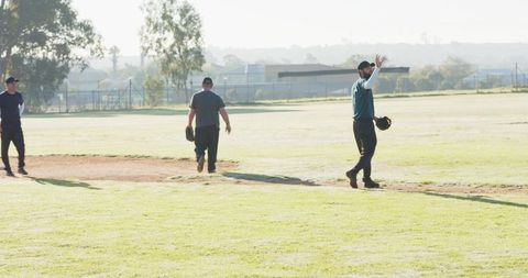Diverse teammates practicing baseball on sunny outfield holding gloves and waving caps
