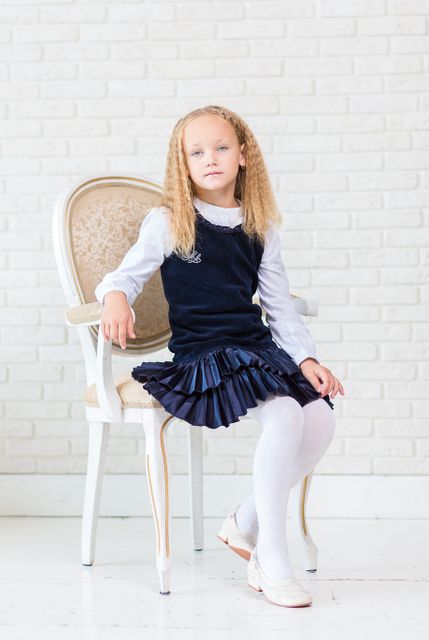A young school girl sitting on an elegant white chair wearing a blue school uniform with pleated skirt. She is looking at the camera with a composed expression, her blonde hair styled in soft waves. The background is a light brick wall, giving a bright and clean atmosphere. This image can be useful for educational content, school promotions, children’s fashion showcases, or childhood education themes.