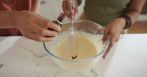 Two women whisking batter pouring food coloring into glass bowl on countertop