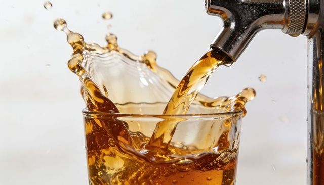 Pouring amber beer from stainless tap creating dramatic crown splash over clear glass