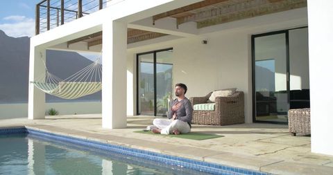 Indian man meditating on yoga mat at poolside patio with hammock and lakeside view