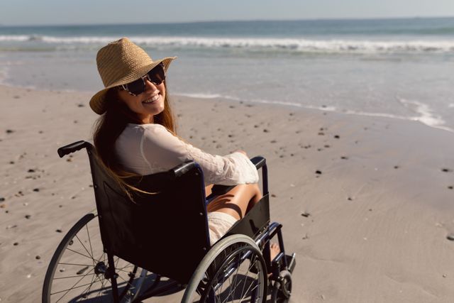 Disabled happy woman in hat and sunglasses sitting on wheelchair at beach in the sunshine