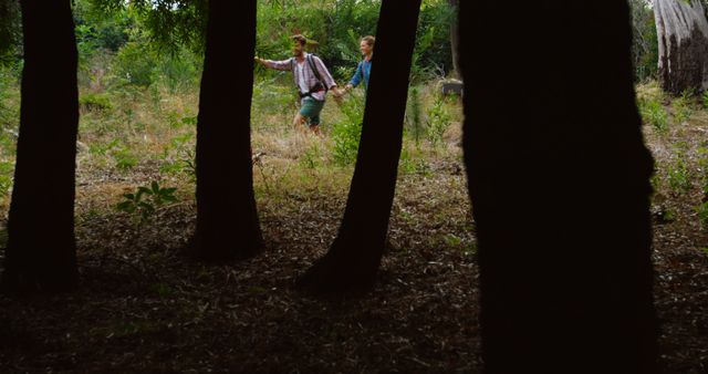 Two young individuals are seen exploring a forest, partially obscured by tree trunks in the foreground. Their adventure through the natural environment suggests a sense of curiosity and discovery.