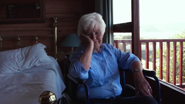 Caucasian elderly woman sitting in wheelchair inside a cozy bedroom, looking pensive by a large window. The image captures personal reflection and solitude, with serene surroundings suggesting home comfort. Use this for content focused on aging, mobility, solitary contemplation, or domestic lifestyle for seniors.